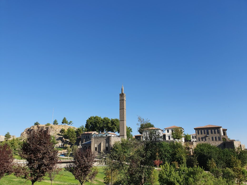 Inner Castle of Amed / Diyarbaki: cluster of historic buildings on an elavation. Buildings are made from black basalt stone, sometimes contrasted with white plastered walls, white mortar, or layers of yellow limestone. Red/orange gable roofs. In the center, a towering black, square minaret with a pointy top. A green grassfield lies before the elavated buildings, but is somewhat obscured by trees of various colors in the foreground. Clear blue skies.
