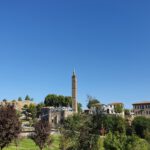 Inner Castle of Amed / Diyarbaki: cluster of historic buildings on an elavation. Buildings are made from black basalt stone, sometimes contrasted with white plastered walls, white mortar, or layers of yellow limestone. Red/orange gable roofs. In the center, a towering black, square minaret with a pointy top. A green grassfield lies before the elavated buildings, but is somewhat obscured by trees of various colors in the foreground. Clear blue skies.