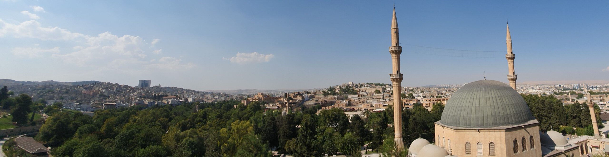 Panorama of the ancient city of Riha/Urfa. Mosque with large dome and two minarets in foreground. Minaret of the 12th century Halil ul-Rahman Mosque is visible. City gardens. Old honey-colored buildings in the background.