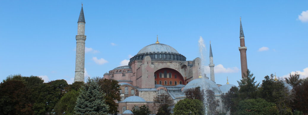 Hagia Sophia: historic structure with large dome, smaller domes, and minarets. Foreground with trees and fountain. Clear blue sky with a few clouds.