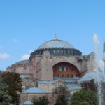 Hagia Sophia: historic structure with large dome, smaller domes, and minarets. Foreground with trees and fountain. Clear blue sky with a few clouds.