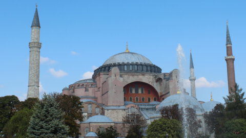 Hagia Sophia: historic structure with large dome, smaller domes, and minarets. Foreground with trees and fountain. Clear blue sky with a few clouds.