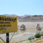 Black text on yellow signboard with the town's name "Hasankeyf". Below it, greetings in various languages: "Hosgeldiniz" (Turkish), "Hûn Bi Xêr Hatin" (Kurdish), "Welcome" (English). Sign is rusty. Background, the Tigris river and the river banks. The Kurdish Ayyubid-era castle is visible. Giant pillars, remnants of an ancient bridge. Some trees. Clear blue sky. Rock formations.
