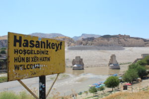 Black text on yellow signboard with the town's name "Hasankeyf". Below it, greetings in various languages: "Hosgeldiniz" (Turkish), "Hûn Bi Xêr Hatin" (Kurdish), "Welcome" (English). Sign is rusty. Background, the Tigris river and the river banks. The Kurdish Ayyubid-era castle is visible. Giant pillars, remnants of an ancient bridge. Some trees. Clear blue sky. Rock formations.