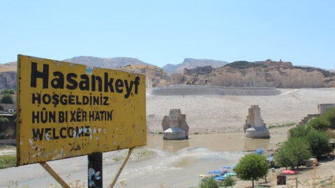 Black text on yellow signboard with the town's name "Hasankeyf". Below it, greetings in various languages: "Hosgeldiniz" (Turkish), "Hûn Bi Xêr Hatin" (Kurdish), "Welcome" (English). Sign is rusty. Background, the Tigris river and the river banks. The Kurdish Ayyubid-era castle is visible. Giant pillars, remnants of an ancient bridge. Some trees. Clear blue sky. Rock formations.