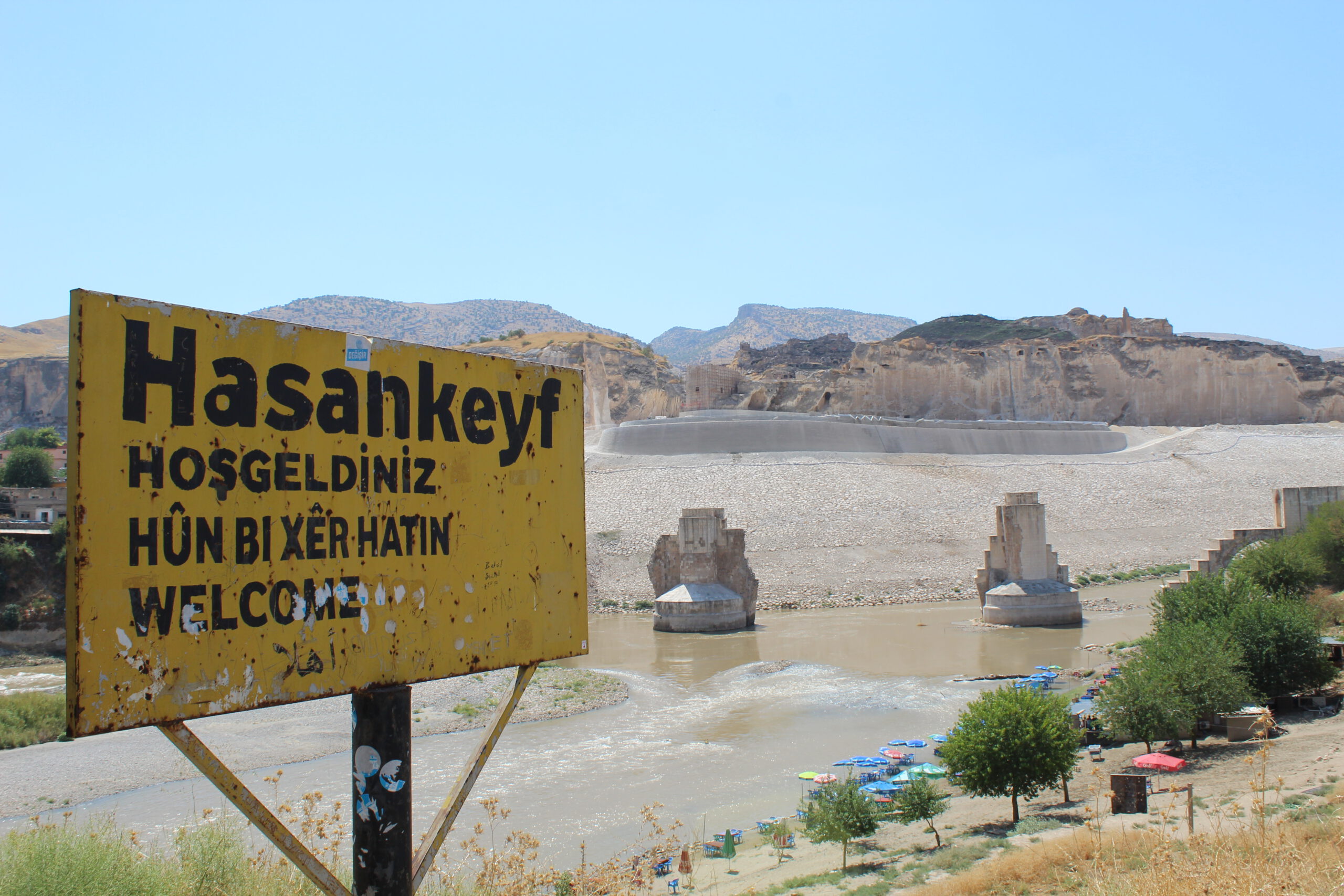 Black text on yellow signboard with the town's name "Hasankeyf". Below it, greetings in various languages: "Hosgeldiniz" (Turkish), "Hûn Bi Xêr Hatin" (Kurdish), "Welcome" (English). Sign is rusty. Background, the Tigris river and the river banks. The Kurdish Ayyubid-era castle is visible. Giant pillars, remnants of an ancient bridge. Some trees. Clear blue sky. Rock formations.